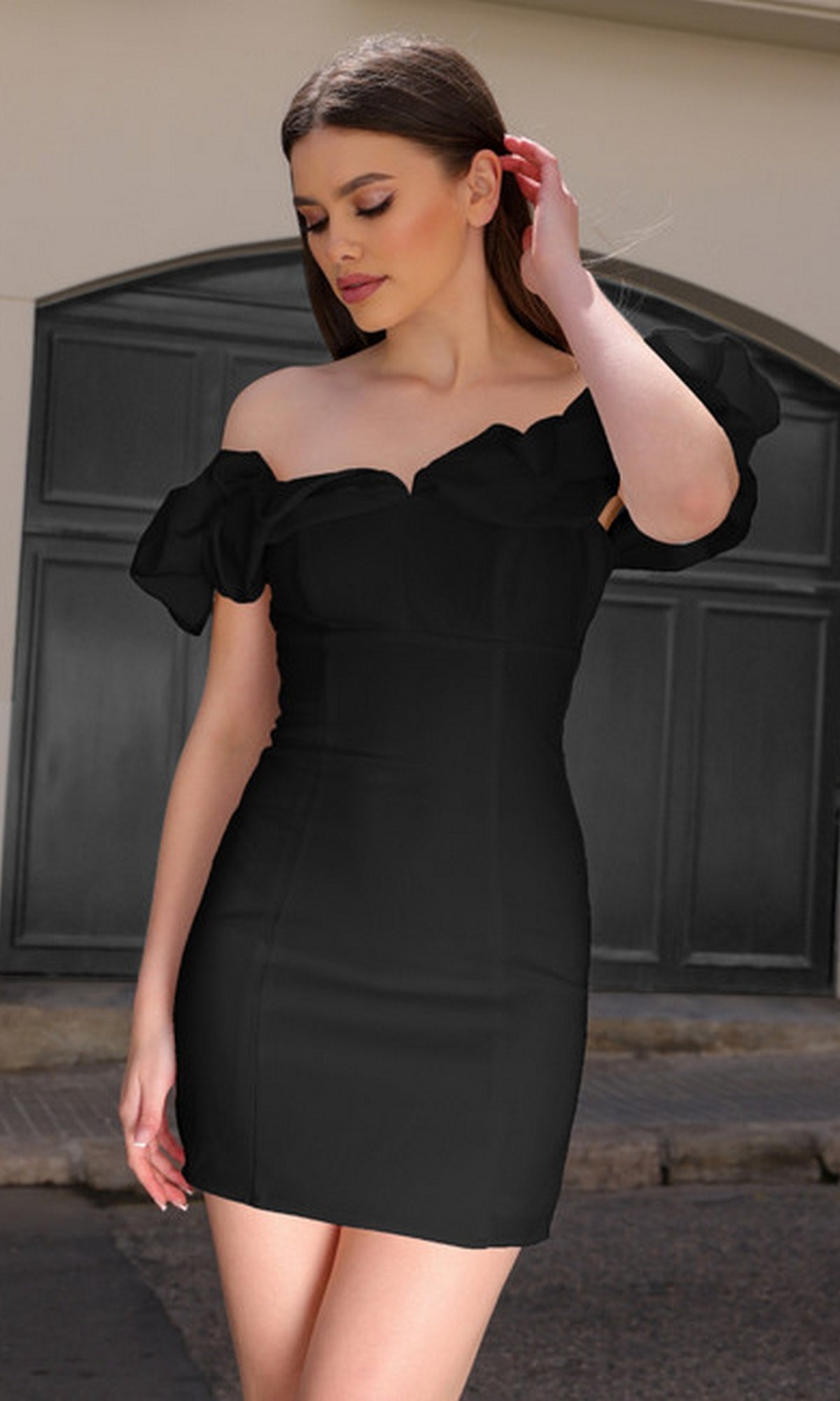 A woman stands outdoors in the Velvi Off-Shoulder Short Homecoming Dress Felicia, its ruffled sleeves and elegant design adding a chic, semi-formal touch as she looks down and gently touches her hair.